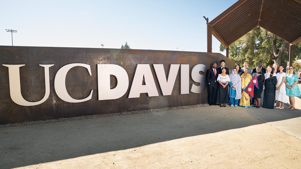 Eleven Humphrey fellows and two program coordinators stand next to a large UC Davis sign on campus. The sign is on a wall of gray concrete with large white letters that read "UC Davis". Humphrey fellows wear a mixture of business and traditional dress.