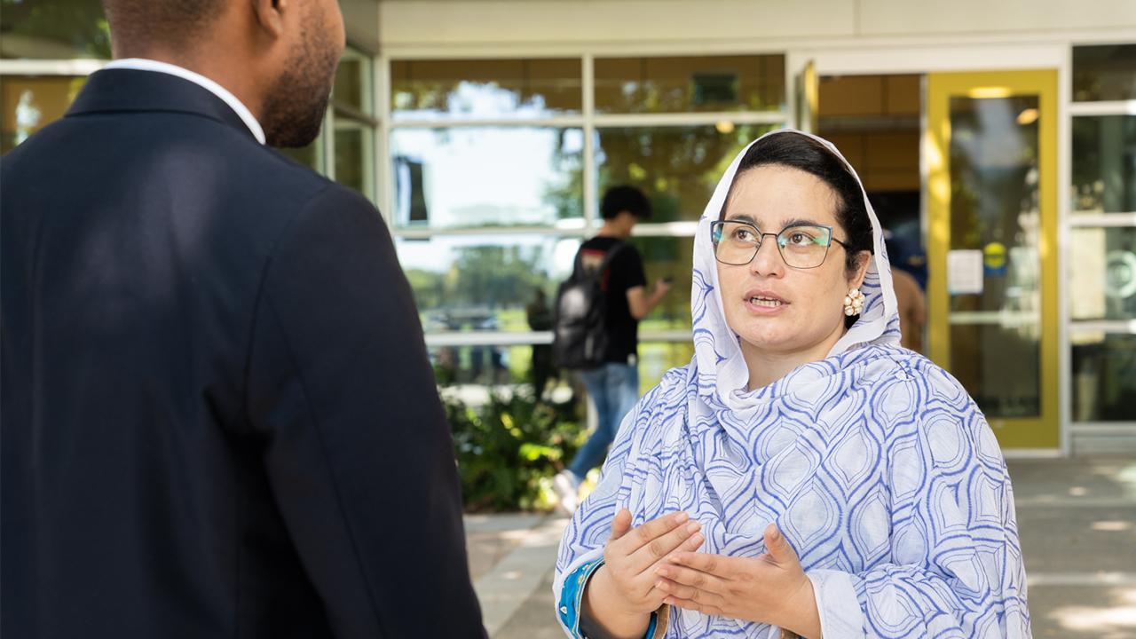 Nadia stands outside the entrance of the International Center and speaks with a colleague. The colleague's back is to us. We see Nadia, wearing a white and blue diamond patterned hajib and large pearl earrings in the shape of a flower, gesture with her hands as she speaks. 
