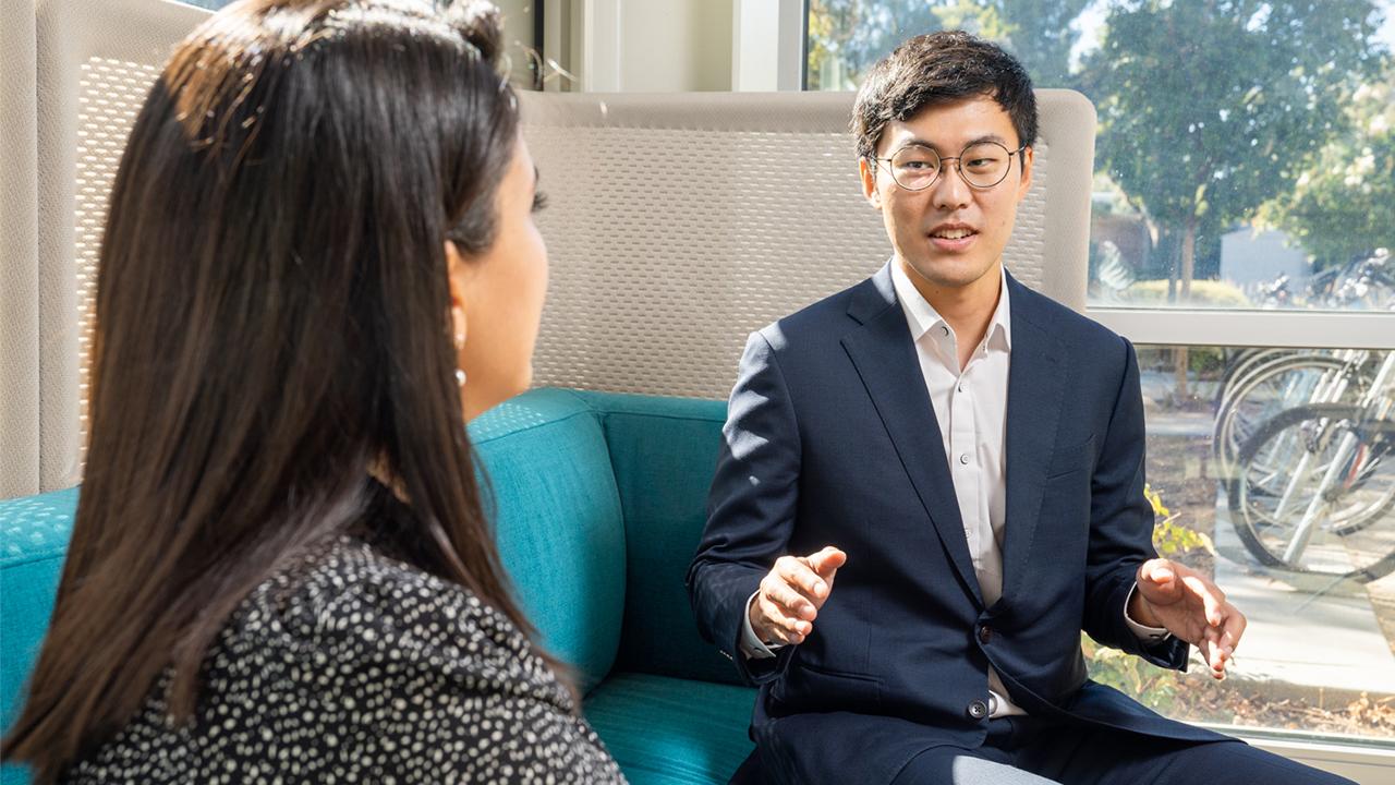 Seungdeok sits inside on a teal couch with windows behind him looking out on a row of parked bicycles. He speaks to a colleague, who is visible from the back.