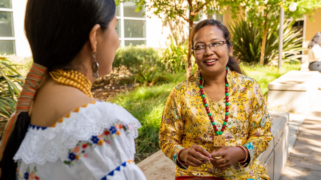 Sushila sits on a bench outside the International Center. She smiles as she speaks with a colleague, whose back it to the camera.