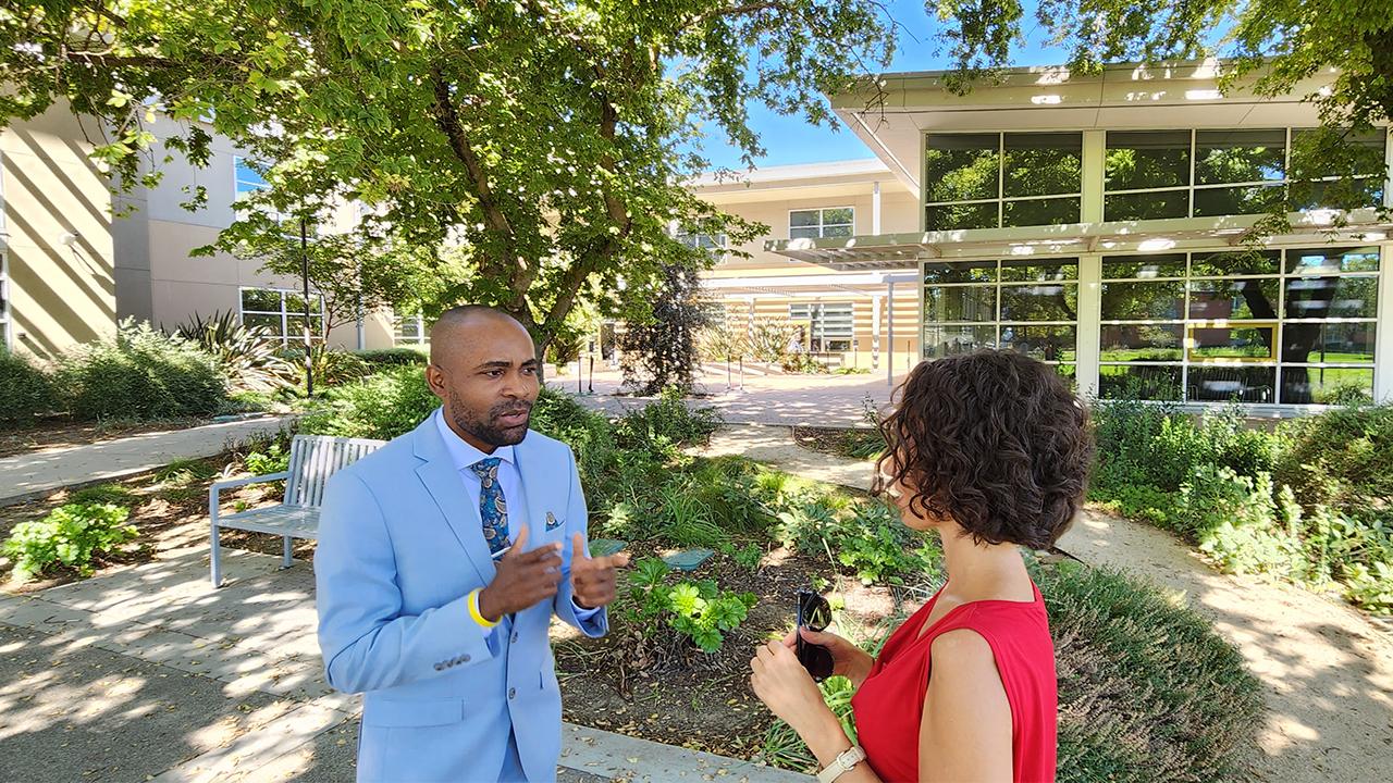 Edu speaks to a woman dressed in a red sleeveless top outside the International Center on the UC Davis campus. They stand in an area shaded by the trees on a sunny day. Edu uses his hands to speak and they are blurred in the photograph. He wears a light blue suit with a matching shirt and blue and yellow paisley tie and pocket square.