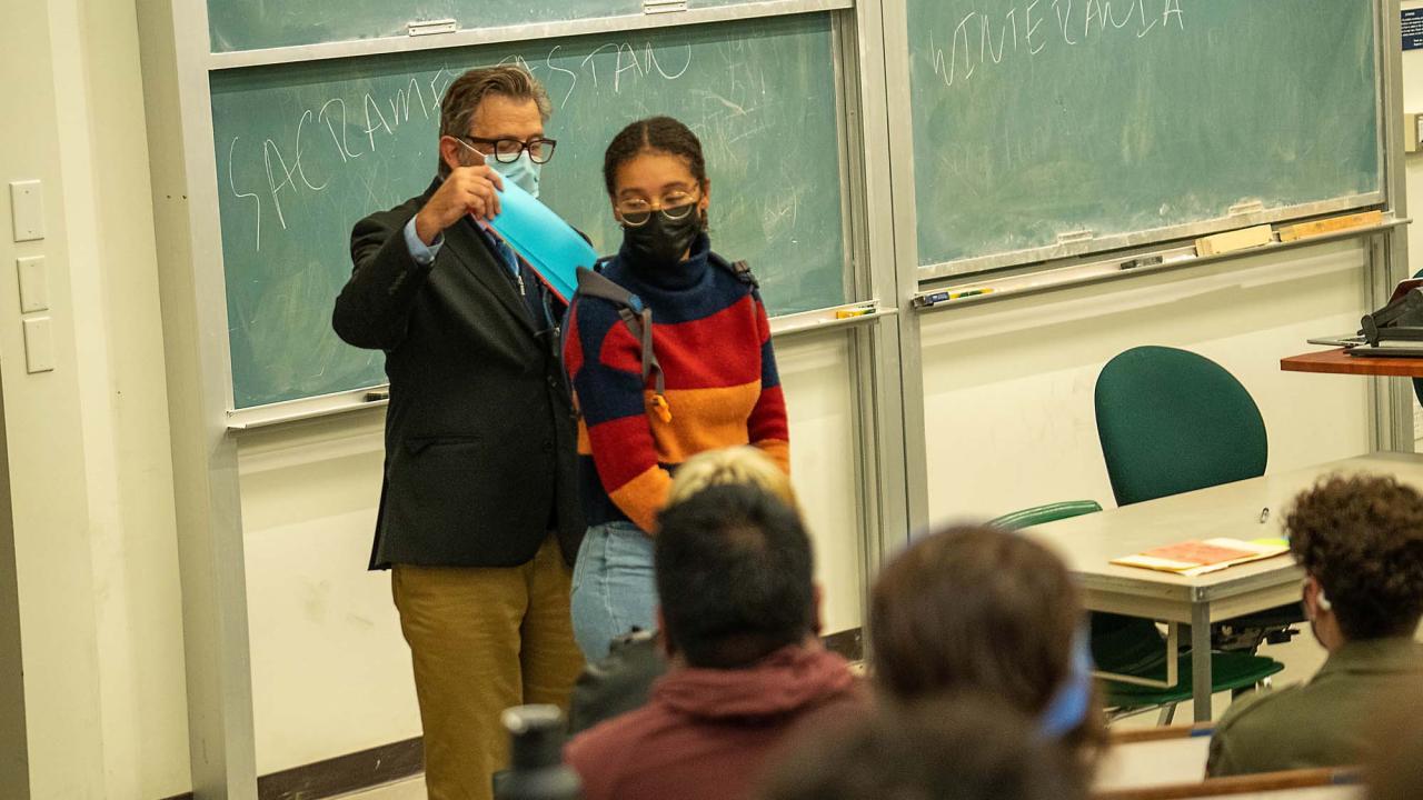 Keith David Watenpaugh stands at the front of a classroom putting a blue paper into a backpack worn by Naomi Danner. Both wear face coverings. Dr. Watenpaugh wears a dark green jacket and khaki pants, and dark rectangular framed glasses. His salt and pepper hair is neatly styled longer on top. Naomi stands in front of him wearing silver round-shaped framed glasses and a turtleneck blocked in navy blue, red, orange and navy blue, and light blue jeans. Her curly black hair is pulled back.