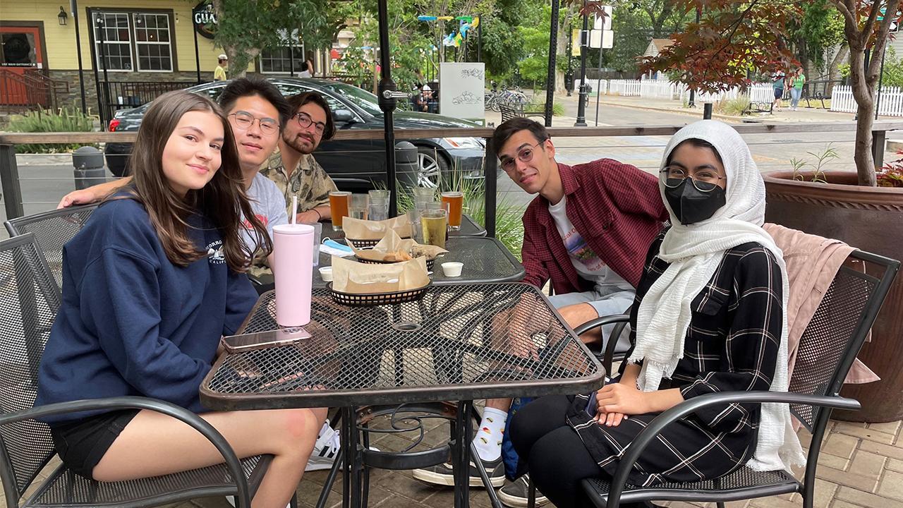 Five students sit around cafe tables outside of a restaurant. They have drinks and food in baskets in front of them. 