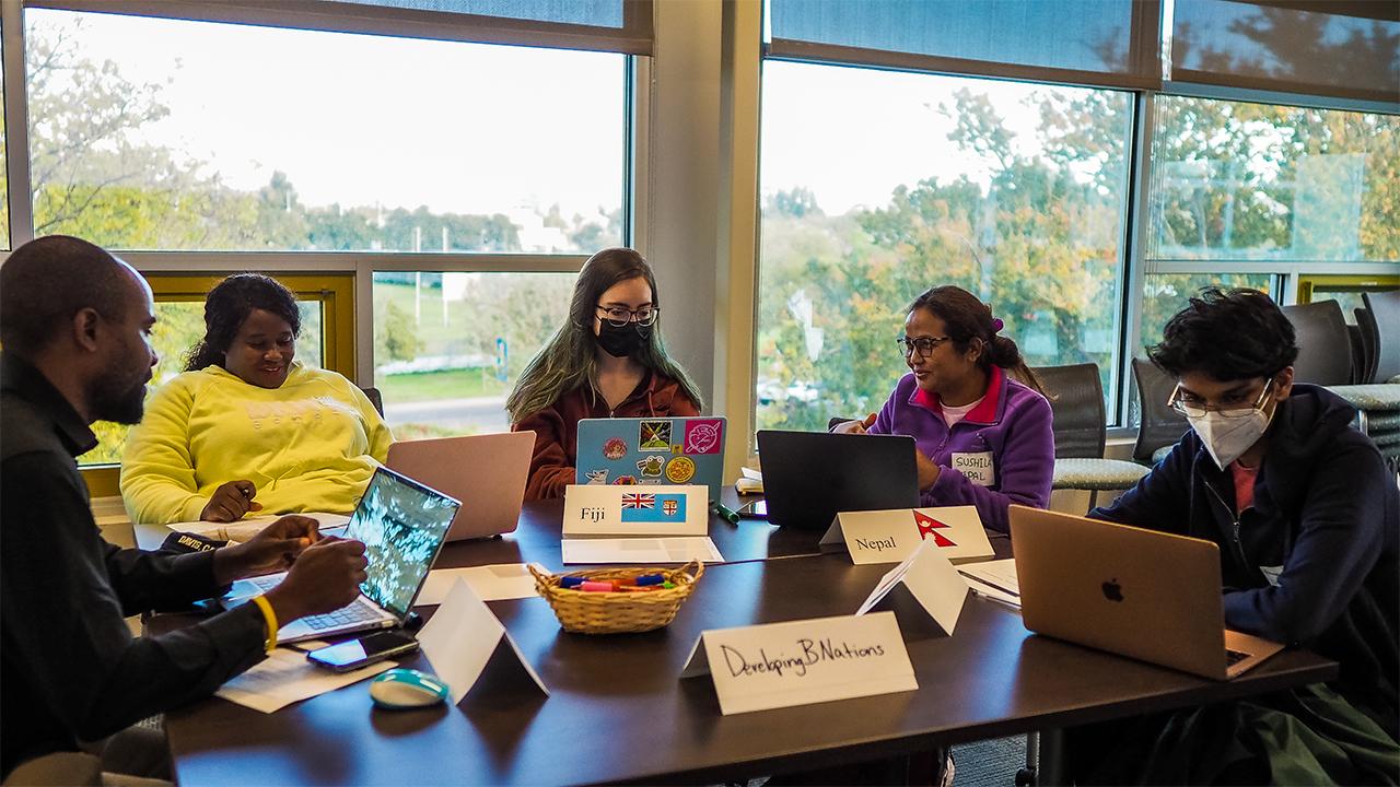 Gathered around a table with windows behind them looking out on trees and a grassy field, Edu Nguema, Rudo Chasi, a female student in a mask, Sushila Thing and a male student in a mask each have a laptop in front of them. Placards on the table identify this group as "Developing B Nations" and some of the countries they represent as Fiji and Nepal. 