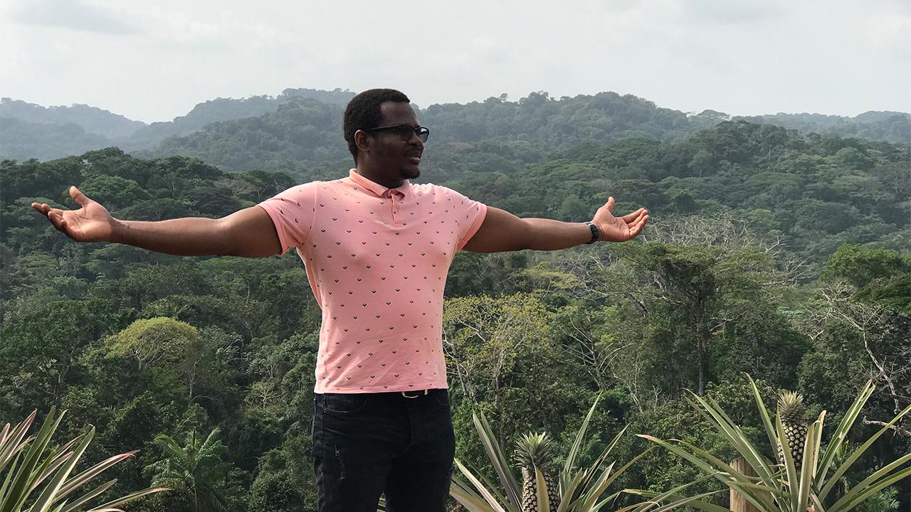 Fabrice stand outside in the nature preserve with lush green trees and plants surrounding him in a valley below. He faces toward the camera but his body and gaze is to the right of the photo as he holds his arms out to his sides as if to take in the majesty of his surroundings.