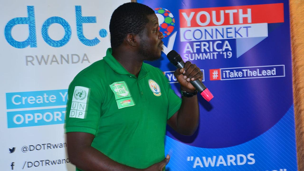 Jireh Bookwa stands before banners for Youth Connekt Africa Summit 2019. He holds a microphone as he speaks to the crowd, and wears a green polo shirt with various patches. His black hair is closely cropped, and he is in profile as he looks to his left.