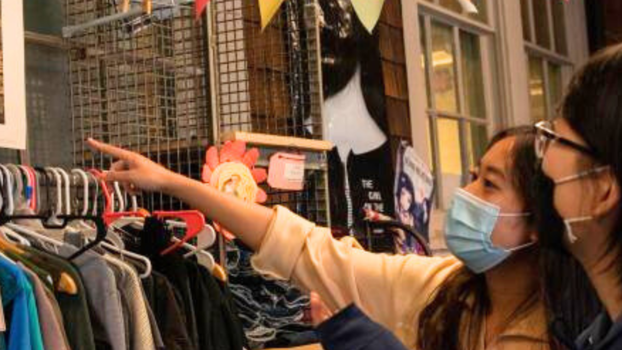Two students wearing face masks stand in front of a rack of clothing and one points to a piece of art on the wall over the racks with a 19th century American or European mother and child.