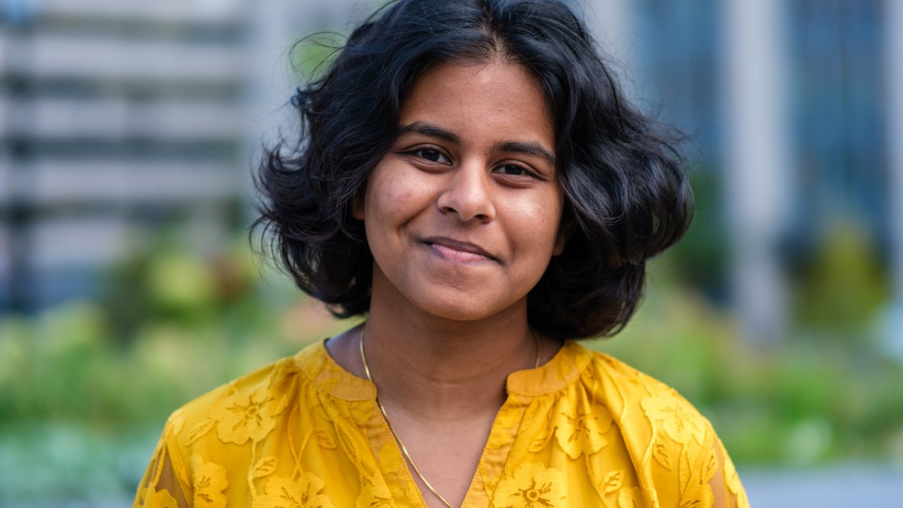 Dheera smiles outside in a yellow blouse with a blurred background of trees and buildings.