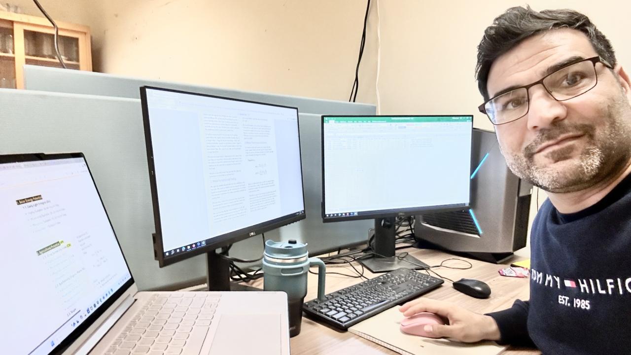 Dr. Ganji sits at a desk surrounded by two monitors and laptop and looks at the camera
