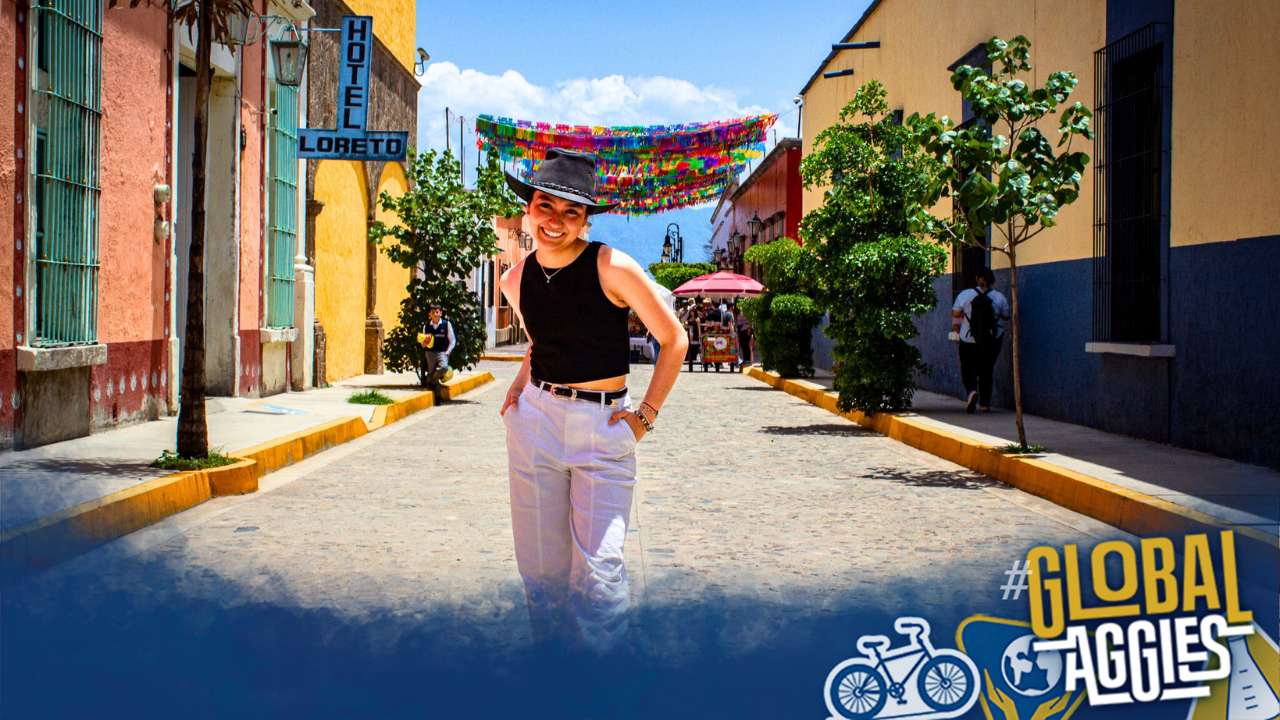 A smiling person walks down a colorful, sunny street with vibrant decorations overhead.