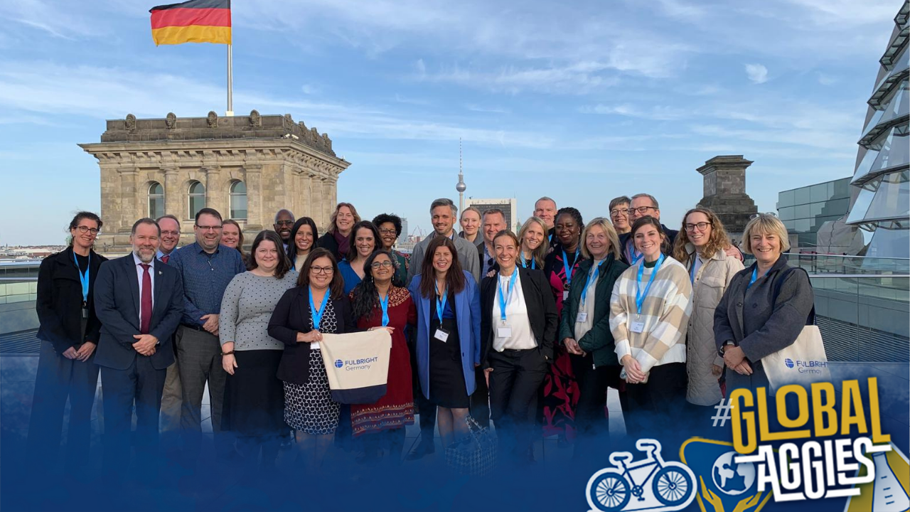 A group of people smile at the camera with a German flag flying in the background. A Global Aggies overlay covers the bottom of the photo.