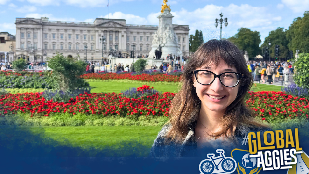 A woman with glasses smiles in front of Buckingham Palace, surrounded by vibrant flower beds.