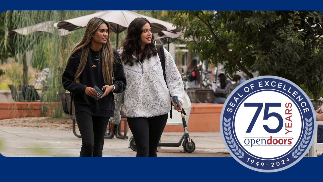 Two students walking on the UC Davis campus with an round seal that says Seal of Excellence 75 Years Open Doors 1949-2024