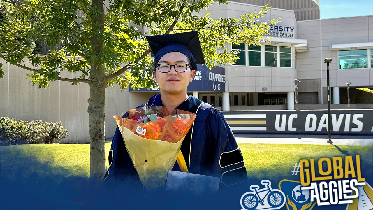 Jeff wearing a graduation cap and gown, holding flowers, and standing in front of a UC Davis sign.