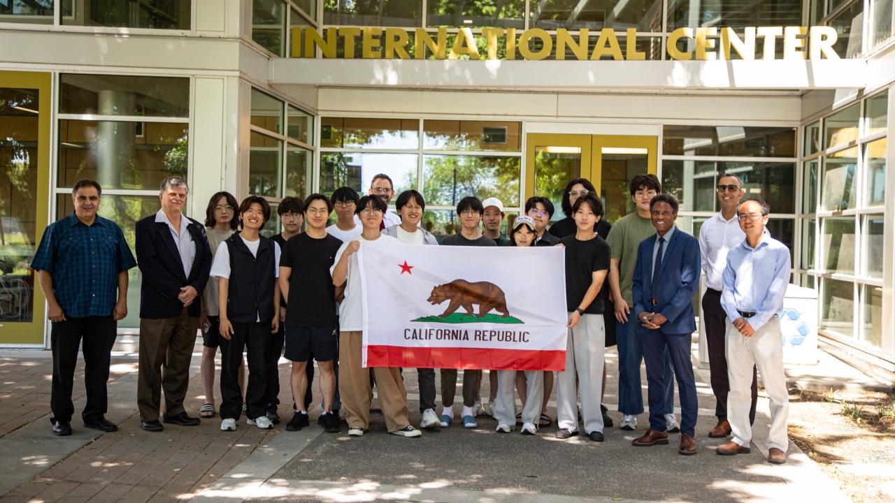 A group of students from Korea University and UC Davis faculty and staff hold up a California flag in front of the UC Davis International Center