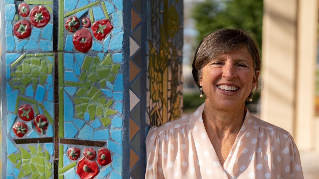 Wolf Prize recipient Pam Ronald outside Robbins Hall, leaning against pillar decorated with a ceramic mosaic mural. Pam has short light brown hair and wears a light pink blouse with cream polka dots.