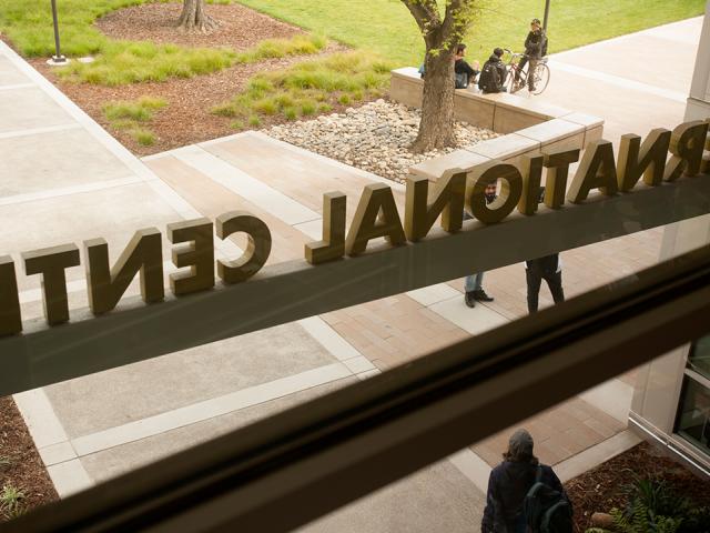 A photo from inside the International Center on the UC Davis campus looking out the main entrance window, taken on a fall day. The International Center sign is backwards from this vantage point looking out as a student wearing a hat, blue jacket and backpack walks away from the doors. Three other students, one sitting on a bike and two others sitting, chat under a tree outside. A student in a pink sweater rides her bike away from the building.