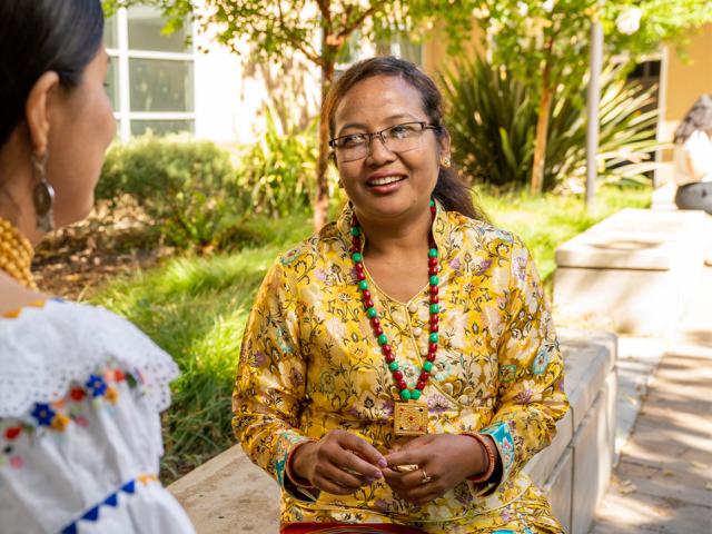 Sushila sits on a bench outside the International Center. She smiles as she speaks with a colleague, whose back it to the camera.