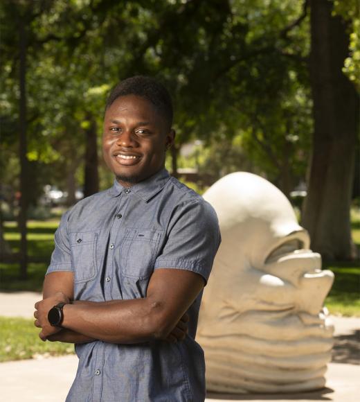 Young man smiling with arms crossed by large white abstract head sculpture on tree-lined campus
