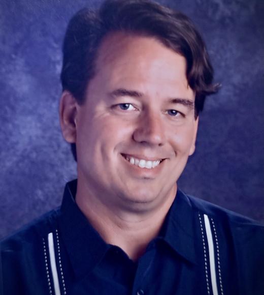 Headshot of a miling man with wavy hair wearing a dark shirt
