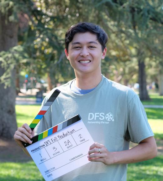 Ryley smiles outside holding a movie clap board wearing a sage green shirt with the letters "DFS."