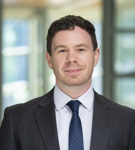 Professional headshot of a man in dark suit and tie, soft smile, blurred office background