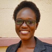 Joanita Ayenyo smiles at the camera wearing a tweed blazer and dark framed rectangular glasses. Her hair is short and she stands outside in front of a cream colored wall with pink trim.