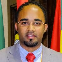 Headshot of Giliardo Lopes Nascimento. He stands in front of flags with red, yellow, and green, wearing a light gray suit, light blue collared shirt, and red tie. His hair is short, he wears frameless rectangular glasses and has a beard on his chin.