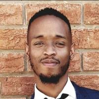 Headshot of Khwezi Mohoang taken outdoors against a brick wall as he smiles slightly to the camera. He wears a navy blue blazer, white collared shirt and black tie. His groomed hair is short on the sides and longer on the top and he wears a short beard. 