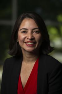 Ana Lucia Arteaga smiles in this 2021 headshot. Her short brown hair is styled in a bob. She wears small gold earrings and a black blazer over a red V-neck blouse.