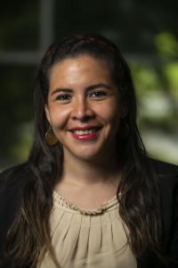 Ana Dominguez smiles at the camera in this 2021 headshot. Her long brown hair hangs over her shoulders. She wears a thin tortoiseshell headband, large gold medallion earrings, a black blazer, and ruffled taupe blouse.