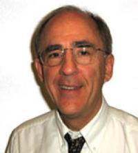Gerald Kost smiles, wearing a white collared shirt and dark tie. He has wire framed glasses. 