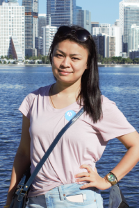 Konsam poses with her hand on her hip in a pink t shirt and jeans with water and a city skyline in the background.