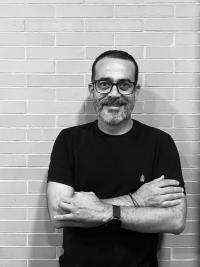 Man with glasses and a beard smiling, arms crossed against a light brick wall.