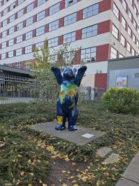 A standing bear statue painted with a world map and a UN Sustainable Development Goals badge is situated on a platform in an outdoor garden area, framed by urban surroundings and a large, red-and-white building.