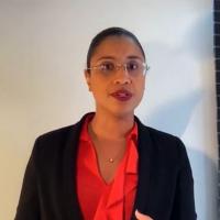 Claudia Duarte Silva Gomes speaks in front of a white wall indoors. Her black hair is pulled back and she wears frameless rectangular glasses, a red blouse, black blazer, and a delicate gold necklace.