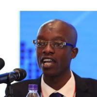 Chipampa Chola speaks at a microphone with a blue background. He wears a dark suit and tie, a white collared shirt, and rectangular glasses.