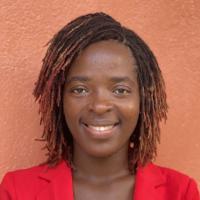 Headshot of Bridget Kowo taken in natural light as she smiles at the camera and stands in front of a textured wall painted terracotta red. Her blazer is candy apple red, and her shoulder length hair is worn in locs, darker at the top and lighter at the ends.