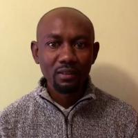 Tangeni Shituleipo Hangula stands before a tan wall indoors. He wears a gray knit sweater with a zipper. His hair is cropped short and he has a short goatee.