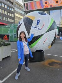 A woman wearing a blue coat and pink shirt stands next to a large soccer ball sculpture with colorful patterns and the Euro 2024 Germany emblem, in front of modern city buildings.