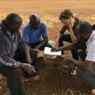 Four people seated outdoors, leaning over papers on dry reddish-brown soil
