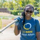 Larissa Kambani stands in the sun holding a gardening tool over her right shoulder. She smiles broadly at the camera, her black hair worn in long braids some of which are pulled back for the hard work she's doing. She wearing black rectangular sunglasses, a heather blue T-shirt with a circle logo that says "UC Davis Global Affairs" and a blue work glove on her right hand. She stands in a park with trees, plants, and a neighborhood street visible behind her.