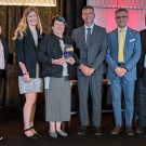 Six professionals stand on a stage, smiling. The woman in the center holds an award.