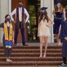 Six students wear commencement regalia and stand socially distanced wearing face masks on the steps of Mrak Hall on the UC Davis campus.