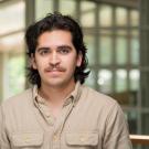 Giovani has long wavy, dark hair he wears styled pushed back off his face. He has a short mustache and wears a beige button-up collared shirt.