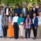 The 24 Fellows stand in two rows, with the back row elevated, under the shade of a large tree. Fellows wear a combination of business casual and cultural attire.
