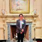 Ana stands in front of an ornate white fireplace and mantel. Over the fireplace is a classic painting and on either side of it are two flags: On the left is the American flag and on the right is the flag of the Office of the President of the United States of America.