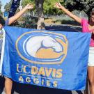 Kara and Rosani each stand on either side of the blue flag they are holding. They smile broadly and each extend an arm up in a flourish. The flag has the UC Davis mustang image and underneath in yellow it says "UC Davis Aggies."