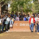 The cohort of 24 Mandela Washington Fellows gathers around a UC Davis sign on a short brick wall on the UC Davis campus.