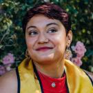 Sarai looks off into the distance. She had short dark hairs and she wears a blue and gold graduation stole over a red top. Her necklace is turquoise and her earrings are gold in a sunburst shapes. She has a gold stud on her nose.